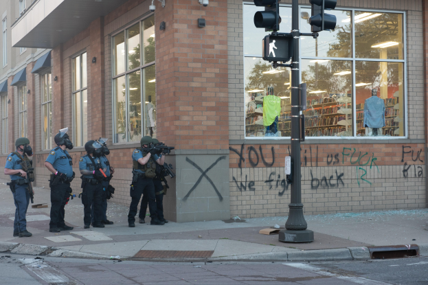 Gruppe von Polizisten in Helmen und taktischer Ausrüstung vor einem Gebäude mit Glasfenstern, bewaffnet, mit einem Verkehrszeichenmast im Vordergrund und Text an der Wand des Gebäudes.