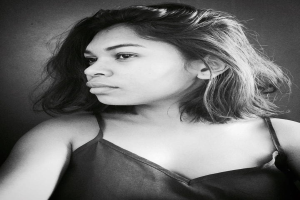 Eine selbstbewusste Frau in einem schwarzen Kleid posiert in einem Schwarz-Weiß-Foto, steht mit einer eleganten Haltung vor einer Wand und trägt einen sleek Bob und dezenten Make-up.