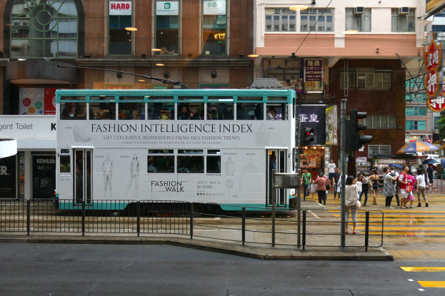 Doppeldeckerbus fährt auf einer Straße mit hohen Gebäuden, Passanten auf dem Gehweg mit Regenschirmen, Ampeln, einem Geländer, Texttafeln, Bäumen und einem klaren blauen Himmel.