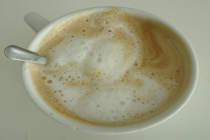 Eine Tasse Cappuccino mit einem Löffel, der an seinem Rand ruht, auf einer weißen Oberfläche gestellt, gefüllt mit einer hellbraunen, cremigen Flüssigkeit.
