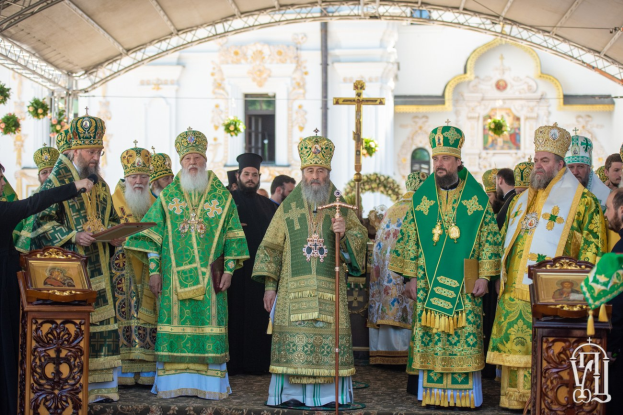 Eine Gruppe von Priestern steht vor einer Kirche während einer religiösen Zeremonie, wobei einer ein Buch und ein Mikrofon hält, während ein Kreuz, Blumen und ein Gebäude im Hintergrund zu sehen sind.