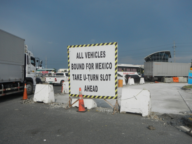 Ein Lastwagen steht auf einem Parkplatz neben einem Schild mit der Aufschrift "Alle Fahrzeuge nach Mexiko", mit verstreuten Kegeln, Brettern und anderen Gegenständen und im Hintergrund Gebäude, Bäume, Pfosten und einen klaren blauen Himmel.