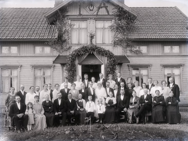 Schwarz-weiß-Foto einer großen Gruppe von Menschen, die vor einem Haus mit Pflanzen posieren.