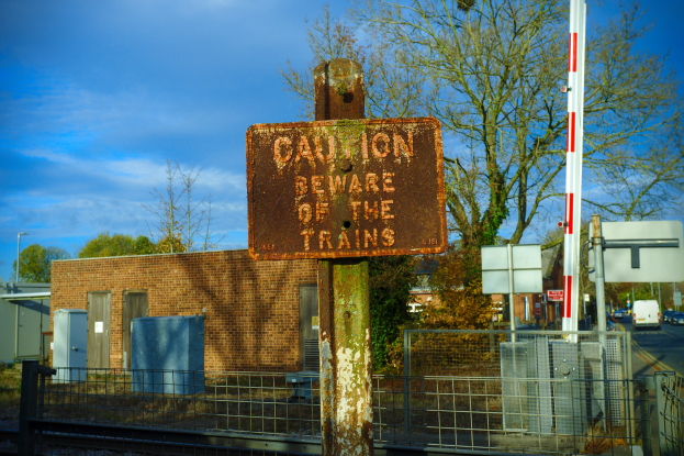 Vorsichtsschild an einem Bahnübergang mit Bäumen, Strommasten, einem Gebäude, Containern, Fahrzeugen auf einer Straße und einem bewölkten Himmel.