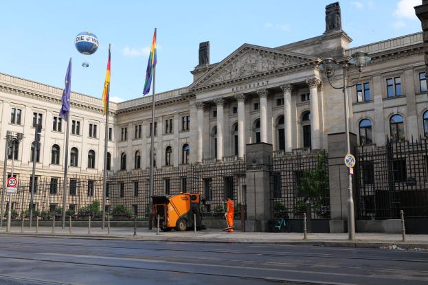 Das Reichstagsgebäude in Berlin, Deutschland, ein großes Gebäude mit zahlreichen Fenstern, umgeben von Straßenlaternen, Fahrzeugen, Menschen und Bäumen, unter einem bewölkten Himmel.