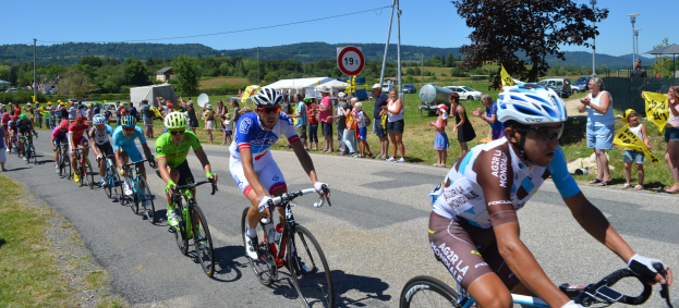 Eine Gruppe von Radfahrern fährt eine Straße entlang, umgeben von Zuschauern, die Fahnen schwenken, einige tragen Helme und Schutzbrillen, im Hintergrund Bäume, Schilder, Fahrzeuge, Zelte, Gebäude, Hügel und einen klaren Himmel.