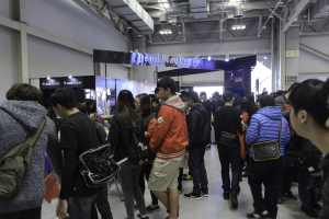 Eine Gruppe von Menschen steht vor einer größeren Menge auf einer Gaming-Messe, wobei einige Besucher Taschen tragen, beleuchtete Texttafeln, Deckenlampen und Rohre im Hintergrund zu sehen sind.