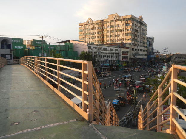 Eine Fußgängerbrücke mit Geländern überspannt eine vielbefahrene Straße mit vielen Fahrzeugen und Fußgängern, mit Gebäuden, Bäumen, Strommasten und einem bewölkten Himmel im Hintergrund.