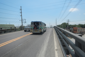 Ein Minibus fährt über eine Brücke mit Geländern, Menschen darin, Strommasten mit Drähten, Bäumen, Gebäuden und einem bewölkten Himmel im Hintergrund.