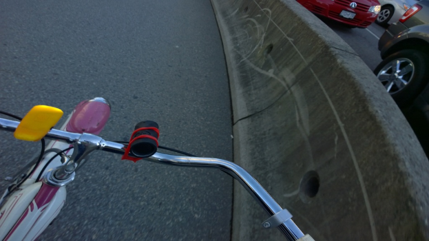 Ein Fahrradlenker ist unten im Bild zu sehen, mit Fahrzeugen, die auf der rechten Seite fahren, und einer Mittellinie.