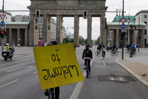 Eine Gruppe von Radfahrern fährt am Brandenburger Tor in Berlin vorbei, einer hält ein gelbes Schild, mit Laternenpfählen, Ampeln, Gebäuden und Bäumen gesäumte Straße.