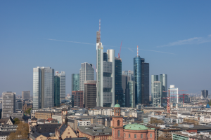 Die Frankfurter Skyline von einem Hügel aus betrachtet, mit einem prominenten zentralen Wolkenkratzer, umgeben von anderen Gebäuden und Bäumen, und dem Himmel im Hintergrund.
