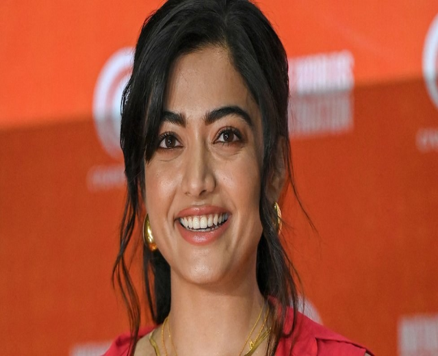 Frau in rotem Kleid mit elegantem Dutt und dezentem Make-up, die in die Kamera lächelt, vor einem gelben Banner mit der Aufschrift "Sonam Kapoors schönste Looks vom Cannes Film Festival."
