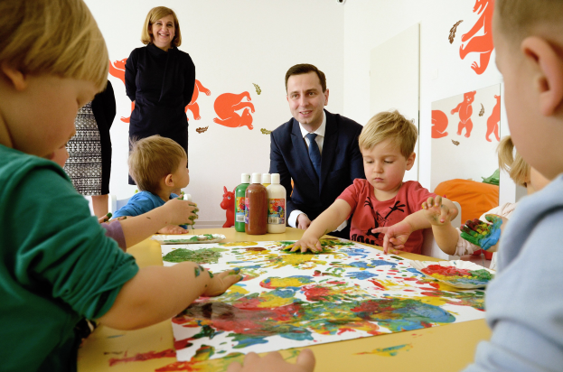 Gruppe von Kindern bei einer Malaktivität um einen Tisch mit farbigen Handabdrücken, Papieren und Flaschen, während eine Frau im Hintergrund mit Kunstwerken an der Wand lächelt.
