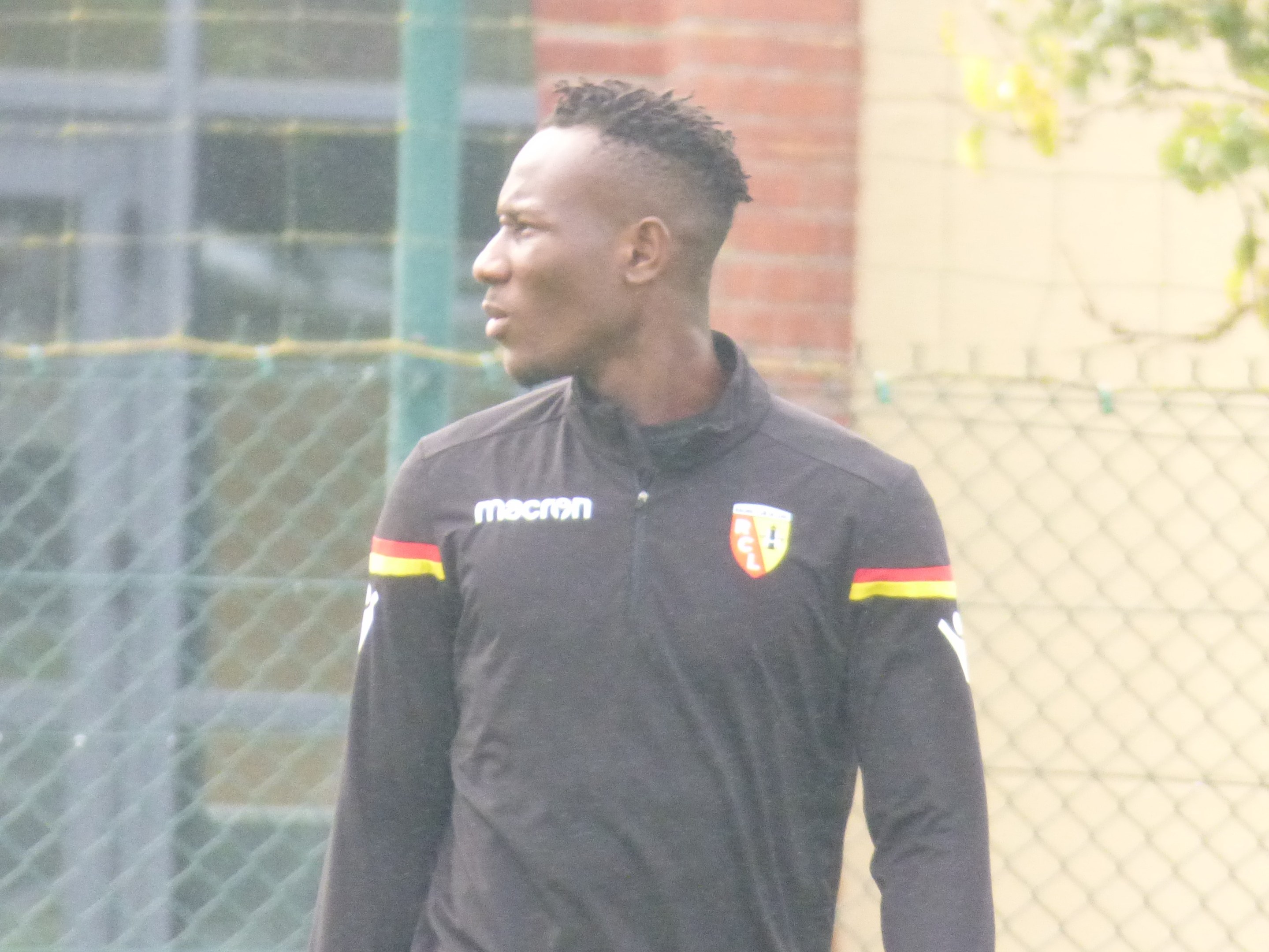 Ein Mann in einer schwarzen Jacke steht auf einem Fussballfeld, umgeben von einem Zaun und einem Baum, und sieht konzentriert während eines Trainings.