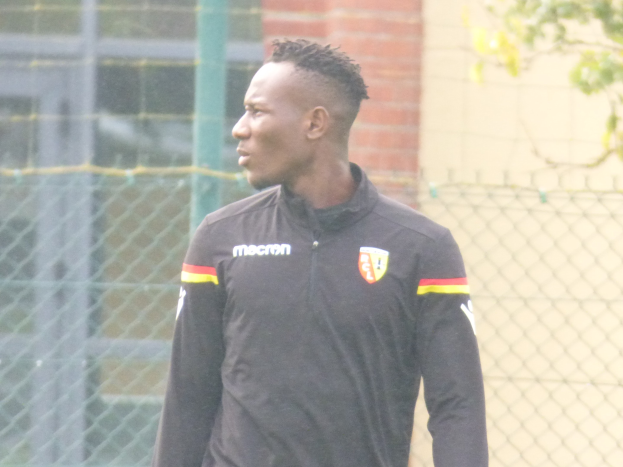 Ein Mann in einer schwarzen Jacke steht auf einem Fussballfeld, umgeben von einem Zaun und einem Baum, und sieht konzentriert während eines Trainings.