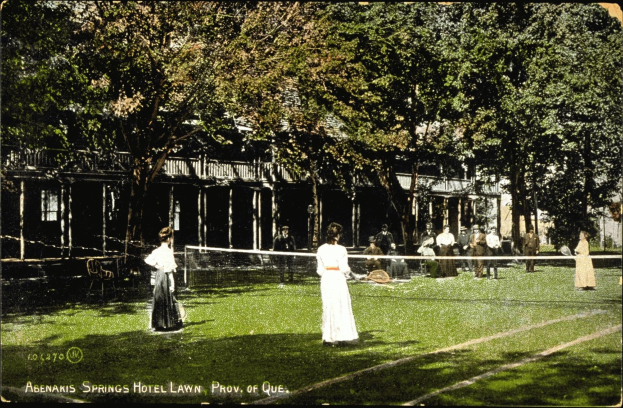 Schwarz-weißes Foto einer Gruppe von Menschen, die auf dem Rasen des Abenakis Springs Hotels in Provo, Quebec, Tennis spielen, mit einem Netz, Stühlen, Bäumen und einem Gebäude im Hintergrund.