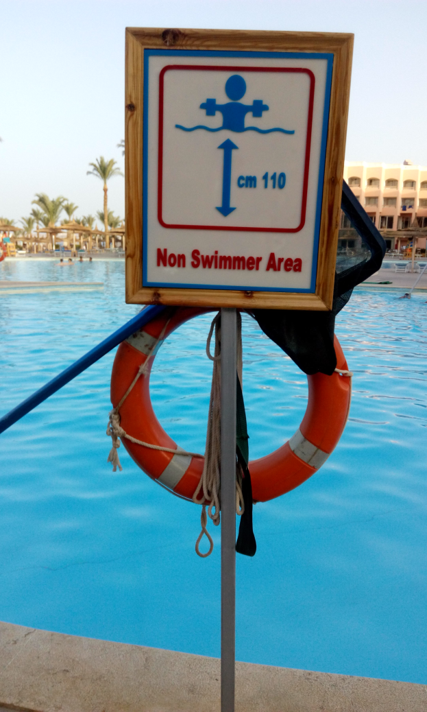 Ein Schwimmbad mit einem "Kein Schwimmer-Bereich"-Schild und einem Rettungsring an einem Pfahl, mit Bäumen, Gebäuden und einem klaren blauen Himmel im Hintergrund.