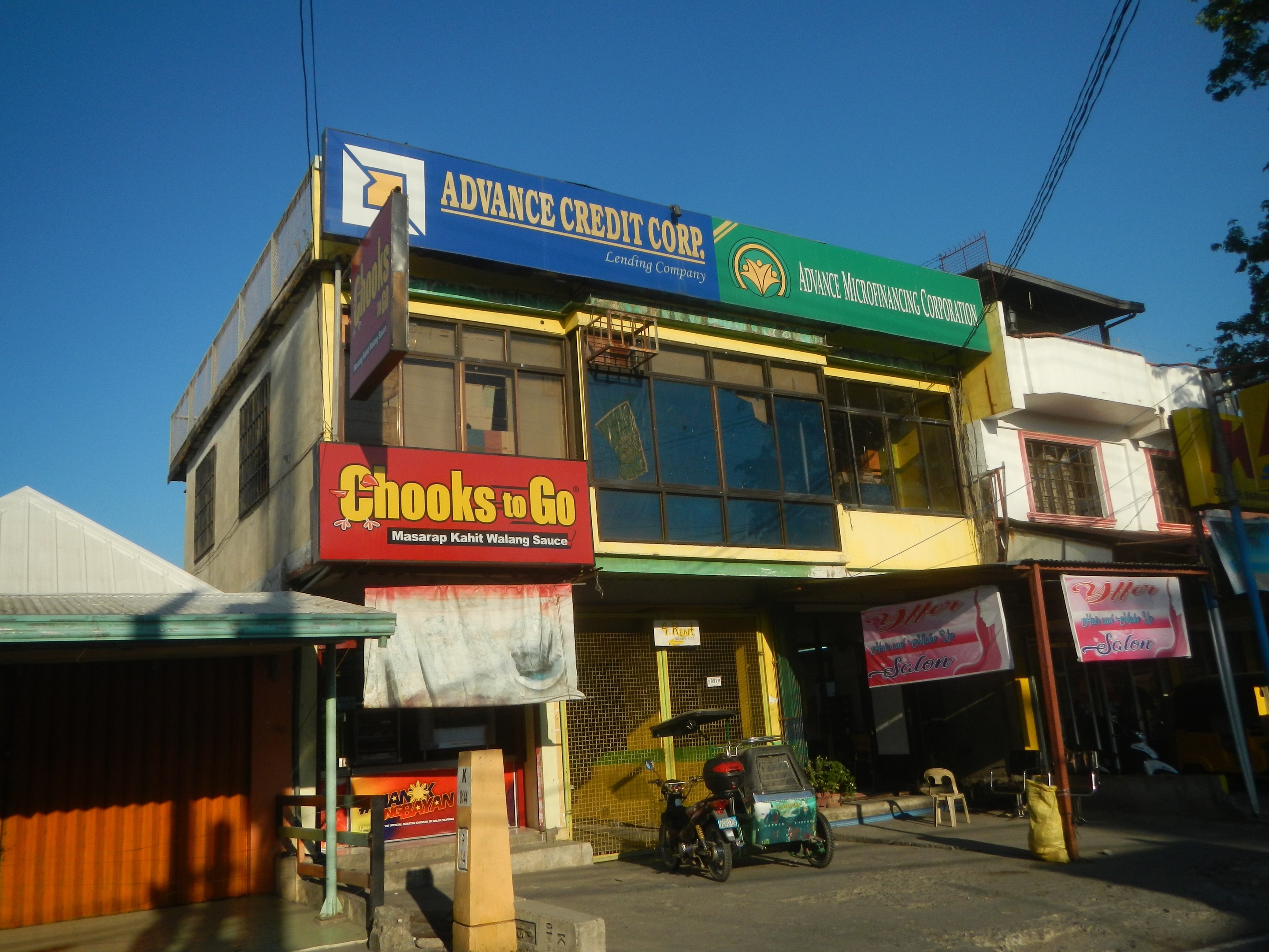 Ein Geschäftsgebäude mit einer Tafel, auf der "Advance Credit Corp" steht, neben einer Straße mit Fahrzeugen, umgeben von Werbetafeln, Strommasten, Kabeln, Bäumen und einem klaren blauen Himmel.