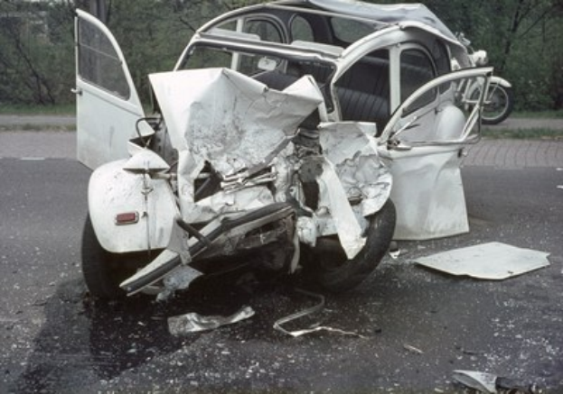 Ein schwer vorne beschädigtes Auto am Straßenrand mit Bäumen und einem Gebäude im Hintergrund und ein weiteres Fahrzeug in der Ferne sichtbar.