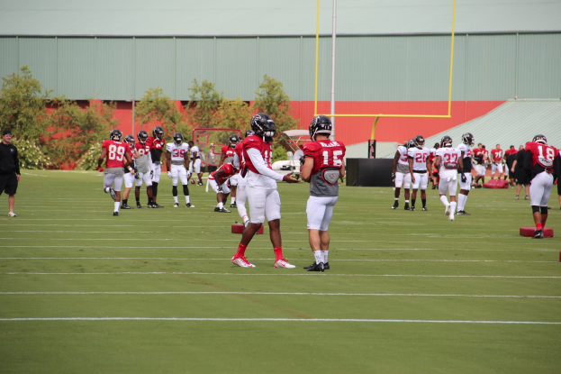 Gruppe von Menschen, die auf einem Rasenfeld Fußball spielen, mit Helmen und Stollenschuhen, umgeben von Bäumen, Pfählen, einer Wand und einem klaren blauen Himmel.