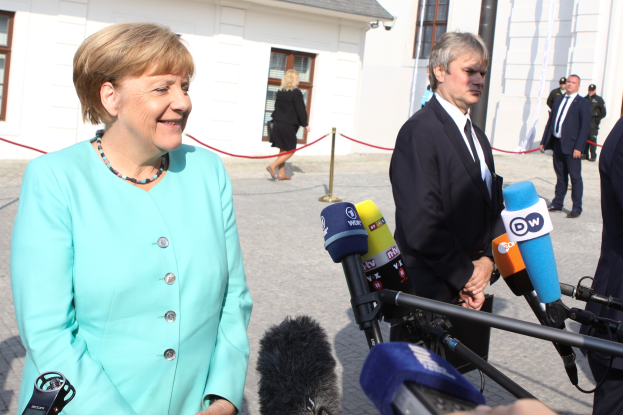 Bundeskanzlerin Angela Merkel beim Sprechen mit Reportern vor einem Gebäude mit Fenstern, umgeben von einer Gruppe von Menschen, einige halten Mikrofone auf Ständern, mit einem Seil, das an einen Pfahl gebunden ist, und einem bewölkten Himmel.