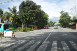 Eine Straßenszene mit Fahrzeugen, einer Zebrastreifen, einem Schild am Boden, Grünfläche, Häusern, Strommasten und einem bewölkten Himmel.