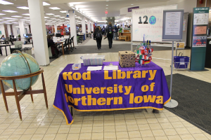 Eine Ausstellung mit einem Tisch, auf dem verschiedene Landesflaggen, eine Box, Papiere und ein Banner liegen, links daneben eine Weltkugel, rechts ein Anschlagbrett mit einigen Menschen daneben und mehrere Tische und Stühle im Hintergrund.