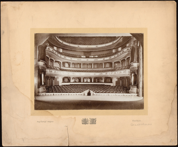 Altes Schwarz-Weiß-Foto des Auditoriums der Royal Albert Hall in London, das die Bühne, Stuhlreihen, Säulen, Geländer und Text mit einem Logo unten zeigt.