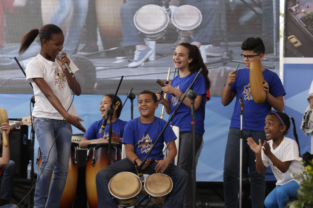 Gruppe von Kindern, die auf einer Bühne Musikinstrumente spielen, mit einer Frau, die ein Mikrofon hält, in der Mitte, umgeben von Mikrofonstativen und einem Banner im Hintergrund, mit Pflanzen auf der rechten Seite.