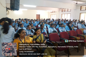 Eine Gruppe von Menschen sitzt in Stühlen in einem Raum mit einer Wand, Fenstern auf der rechten Seite, Deckenleuchten und einem Ventilator und einem Lautsprecher auf der linken Seite, mit Text und einer Karte unten.