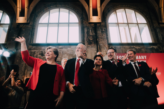 Eine Gruppe von Menschen, wahrscheinlich Labour-Parteimitglieder, die feierlich vor einem Publikum klatschen, mit einem Banner, Glasfenstern und beleuchteter Hintergrund.
