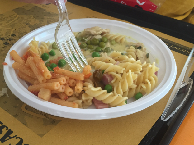 Eine Person hält eine Gabel über eine Schüssel mit Nudeln, Erbsen und Schinken, mit einem Tablett, das einen Löffel und eine Abdeckung enthält.