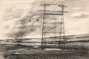 Ein altes Schwarz-Weiß-Foto einer Hochspannungsleitung mit einer Seilbahn im Vordergrund, umgeben von Hügeln und einem bewölkten Himmel im Hintergrund, mit Text unten.