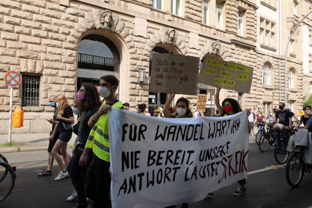 Eine Gruppe von Menschen, die auf einer Demonstration durch die Straße marschieren, einige halten Schilder und andere fahren Fahrräder, im Hintergrund ein Gebäude mit Rundbogenfenstern, Pfeilern und Skulpturen.