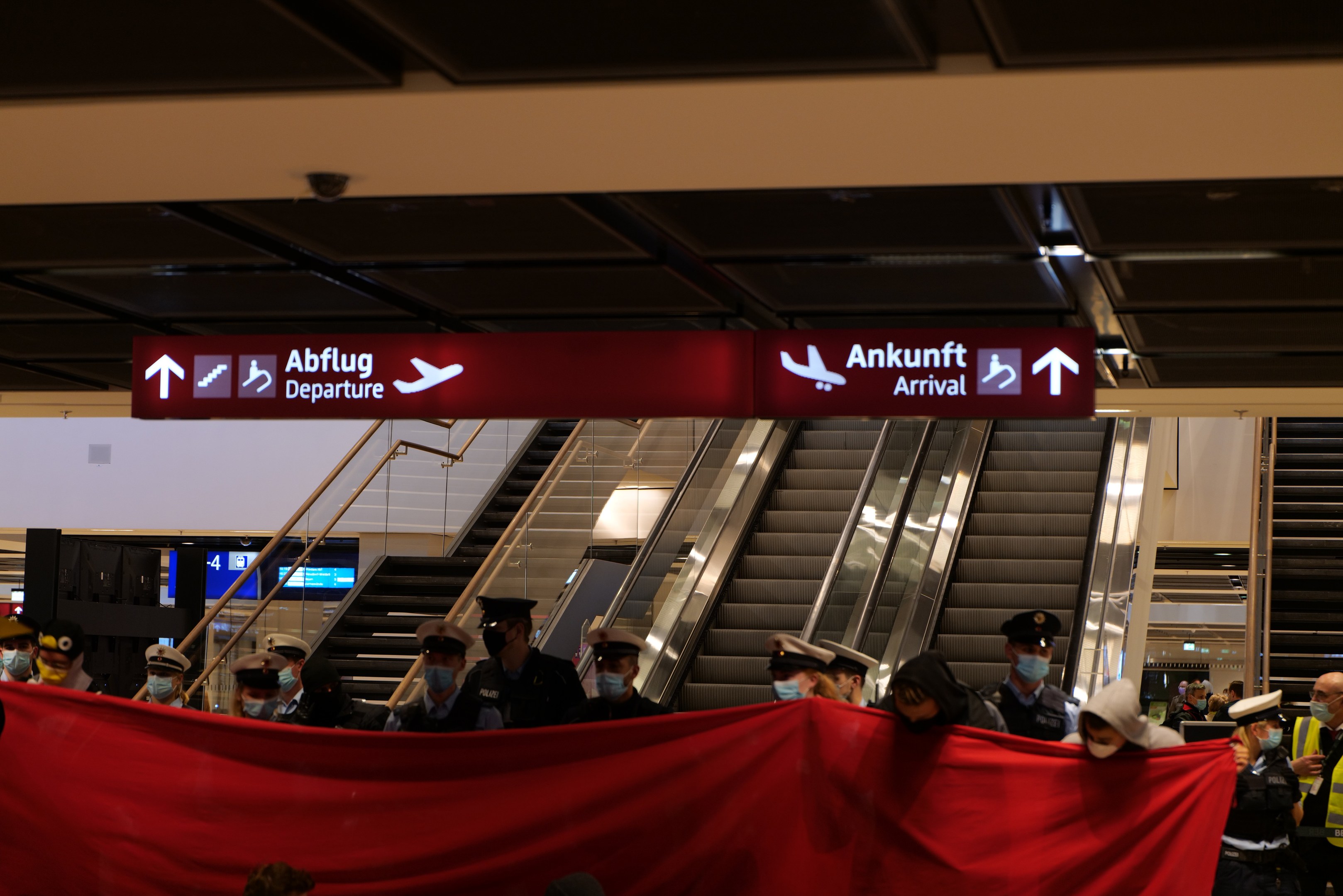 Eine Gruppe von Menschen in Masken und Mützen steht vor einer Rolltreppe und hält ein rotes Tuch und Schilder mit Text darauf in einer Flughafenumgebung mit Deckenbildschirmen und Beleuchtung.