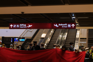 Eine Gruppe von Menschen in Masken und Mützen steht vor einer Rolltreppe und hält ein rotes Tuch und Schilder mit Text darauf in einer Flughafenumgebung mit Deckenbildschirmen und Beleuchtung.