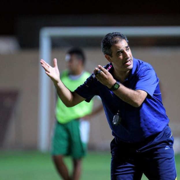 Ein Mann in einem blauen T-Shirt und Hose steht auf einem Fußballfeld und hält ein Mikrofon, während er als Trainer gestikuliert. Im Hintergrund ist eine Person in einem grünen Kleid, ein Tor und eine Wand zu sehen.