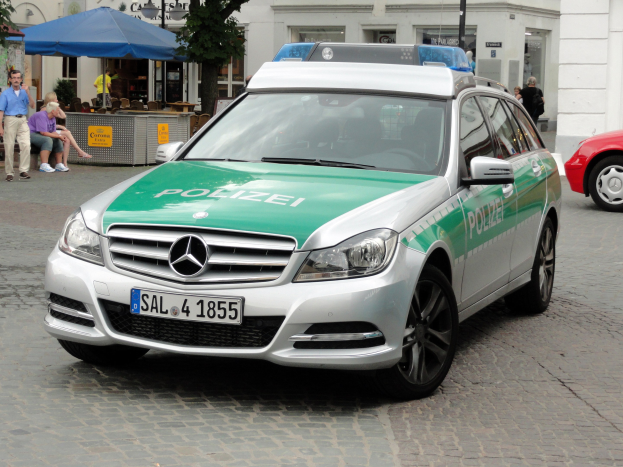 Ein Polizeiwagen neben einem roten Mercedes-Benz C-Klasse W204 auf einer Straße mit Gebäuden, Bäumen und Menschen im Hintergrund geparkt.