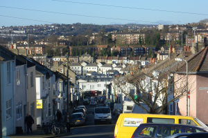 Eine Stadtstraße mit parkenden Autos, Gebäuden mit Fenstern, Bäumen, Laternen und Schildern, mit Menschen auf dem Gehweg und Fahrrädern, die an der Straße geparkt sind, unter einem klaren blauen Himmel.