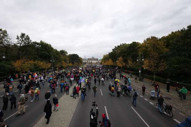 Eine große Gruppe von Menschen marschiert auf einer baumbestandenen Straße mit Laternenmasten, hält Kameras, mit einem Gebäude und einem klaren Himmel im Hintergrund, was auf eine Demonstration in Berlin hindeutet.