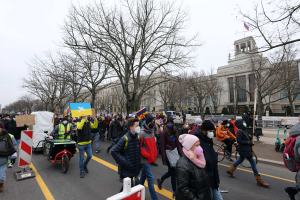 Eine große Gruppe von Menschen nimmt an einer Protestdemo auf einer Straße in Washington, D.C. teil, einige halten Schilder und Transparente, andere fahren Fahrräder, und Schilder, Bäume und ein klarer blauer Himmel im Hintergrund.