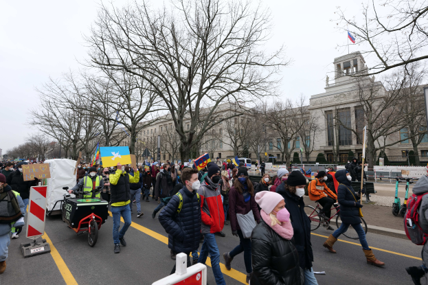 Eine große Gruppe von Menschen nimmt an einer Protestdemo auf einer Straße in Washington, D.C. teil, einige halten Schilder und Transparente, andere fahren Fahrräder, und Schilder, Bäume und ein klarer blauer Himmel im Hintergrund.