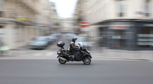 Eine Person in schwarzer Kleidung und Helm auf einem Motorroller die Straße einer Stadt entlangfahren, mit anderen Fahrzeugen, Gebäuden und einem klaren blauen Himmel im Hintergrund.