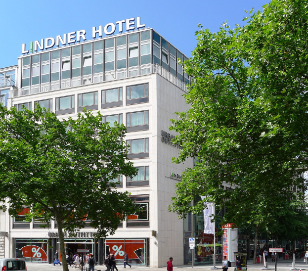 Großes modernes Hotelgebäude mit Glasfenstern und Aufschrift auf der Oberseite, umgeben von Bäumen und einer belebten Straße mit Fußgängern und Fahrzeugen, unter einem sichtbaren Himmel.