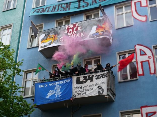 Eine Gruppe von Menschen auf einem Balkon mit Fahnen und Transparenten, mit einem Baum links und Rauch in der Luft, was auf eine Demonstration hindeutet.