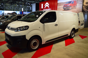 Ein weißer Citroën Berlingo Van wird auf einer Autauschstellung gezeigt, umgeben von schwarzer Schlafzimmer-Möblierung, mit Wänden, die mit Brettern und Lichtern geschmückt sind.