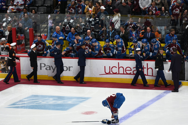 Ein Hockey-Spieler auf dem Eis umgeben von Teamkollegen und Gegenspielern, mit Zuschauern und Arena-Beschilderung im Hintergrund.
