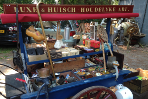 Ein blauer Wagen voller Flaschen und Kisten steht auf einem Schotterweg auf einem Flohmarkt, mit Fahrzeugen, Bäumen und einer Wand im Hintergrund.