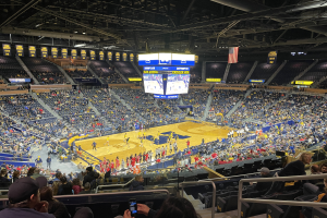 Eine große Arena mit Zuschauern, die ein Basketballspiel verfolgen, wobei einige stehen und andere sitzen, und eine Person mit einem Mobiltelefon in der Hand; die Decke hat Stangen und Lampen, und in der Mitte gibt es Tafeln mit dem Text "Michigan Wolverines Basketball" und einem Bildschirm.